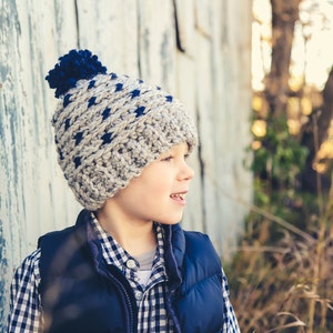 Puede incluir: Un ni&ntilde;o peque&ntilde;o con una camisa a cuadros azules y blancos, un chaleco azul marino y un gorro de punto gris y azul con un pomp&oacute;n. Est&aacute; de pie frente a una pared de madera desgastada.