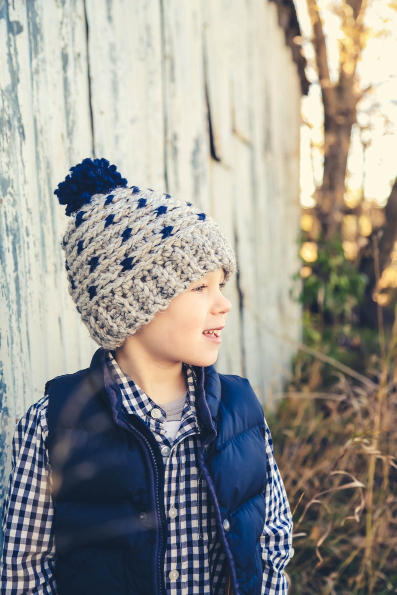 Puede incluir: Un ni&ntilde;o peque&ntilde;o con una camisa a cuadros azules y blancos, un chaleco azul marino y un gorro de punto gris y azul con un pomp&oacute;n. Est&aacute; de pie frente a una pared de madera desgastada.