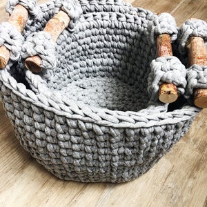 May include: A gray crocheted storage basket with wooden handles. The basket is made of thick yarn and has a woven texture.