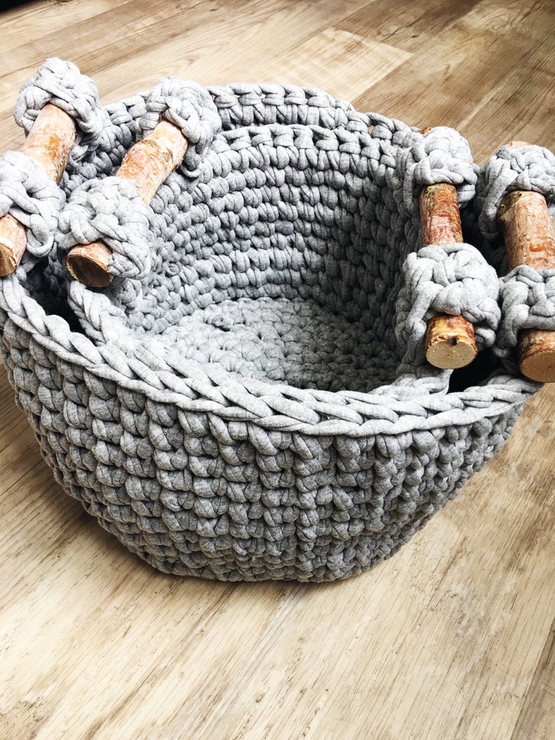 May include: A gray crocheted storage basket with wooden handles. The basket is made of thick yarn and has a woven texture.