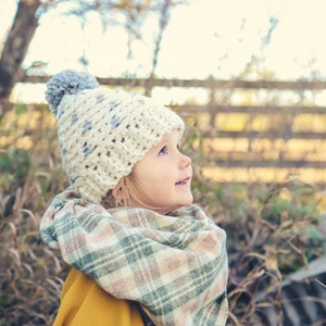 Puede incluir: Una ni&ntilde;a peque&ntilde;a con un gorro de punto blanco y gris con un pomp&oacute;n gris y una bufanda a cuadros. La ni&ntilde;a est&aacute; mirando hacia arriba y sonriendo.
