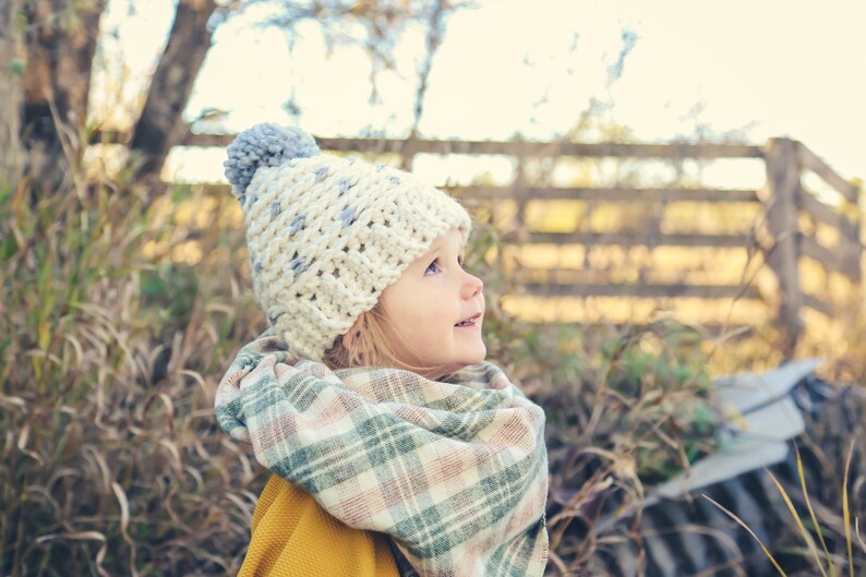 Puede incluir: Una ni&ntilde;a peque&ntilde;a con un gorro de punto blanco y gris con un pomp&oacute;n gris y una bufanda a cuadros. La ni&ntilde;a est&aacute; mirando hacia arriba y sonriendo.