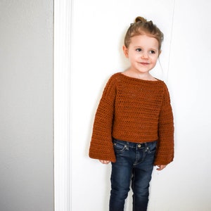 May include: A young person wearing a brown crocheted sweater with bell sleeves and blue jeans.