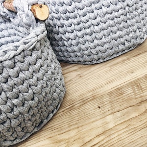 May include: Two gray crocheted storage baskets with wooden handles. The baskets are made of a soft, textured yarn and have a round shape.