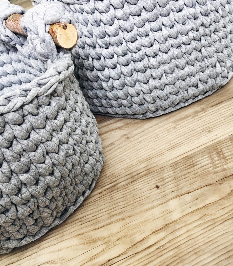May include: Two gray crocheted storage baskets with wooden handles. The baskets are made of a soft, textured yarn and have a round shape.
