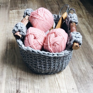 May include: A gray crocheted basket with three pink yarn balls inside. The basket has wooden handles and is sitting on a wooden surface.