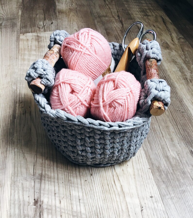 May include: A gray crocheted basket with three pink yarn balls inside. The basket has wooden handles and is sitting on a wooden surface.