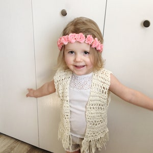 May include: A young girl wearing a white crocheted vest with fringe, a white tank top, and white shorts. She is wearing a pink flower crown.