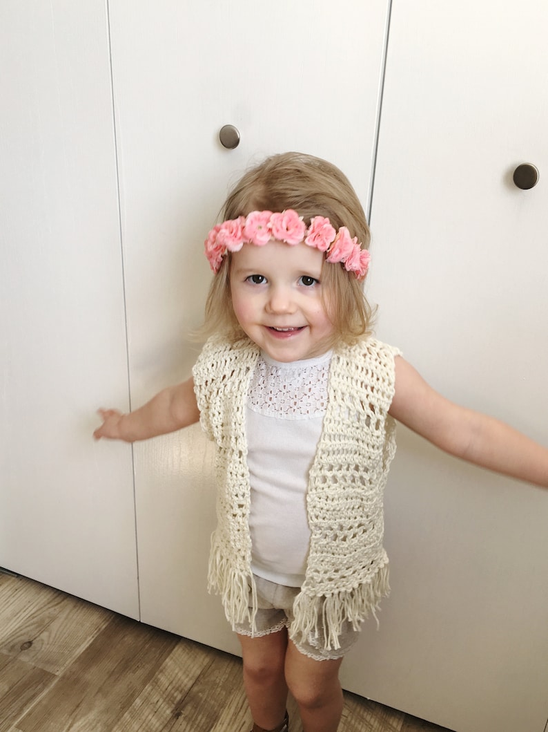 May include: A young girl wearing a white crocheted vest with fringe, a white tank top, and white shorts. She is wearing a pink flower crown.