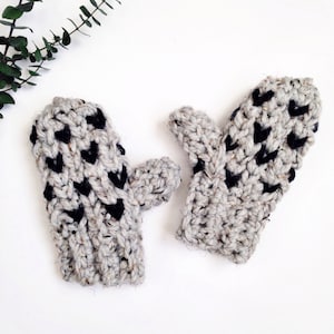 May include: A pair of gray and black crocheted mittens with a heart pattern. The mittens are made with a chunky yarn and have a textured look.