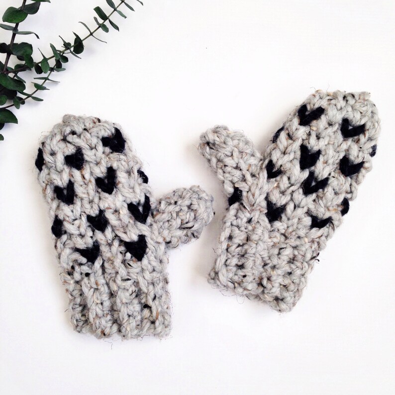 May include: A pair of gray and black crocheted mittens with a heart pattern. The mittens are made with a chunky yarn and have a textured look.