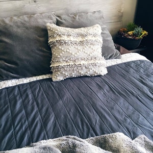 May include: A gray and white crocheted throw pillow with a textured design sits on a gray bed with a gray and white patterned quilt and a gray and white faux fur throw.