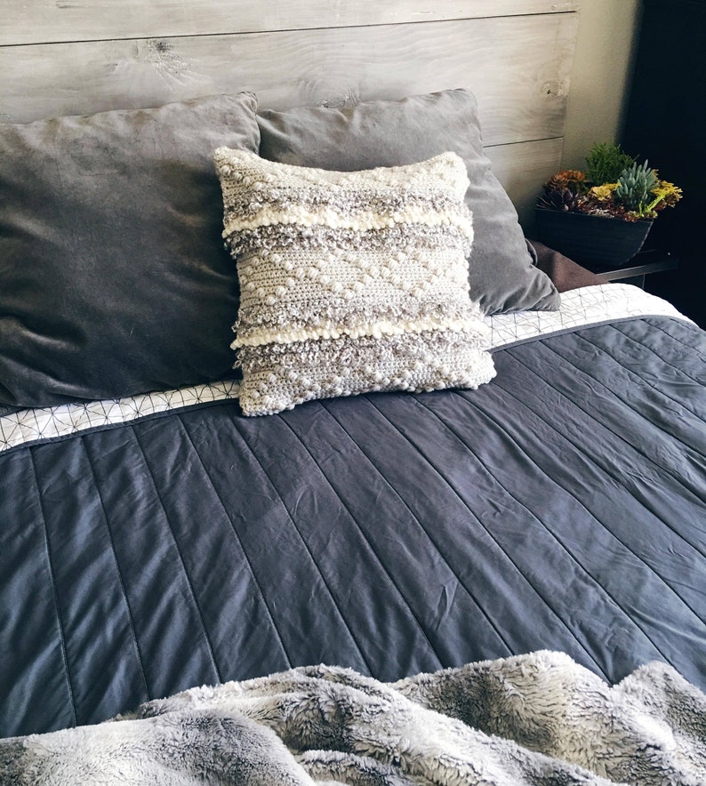 May include: A gray and white crocheted throw pillow with a textured design sits on a gray bed with a gray and white patterned quilt and a gray and white faux fur throw.