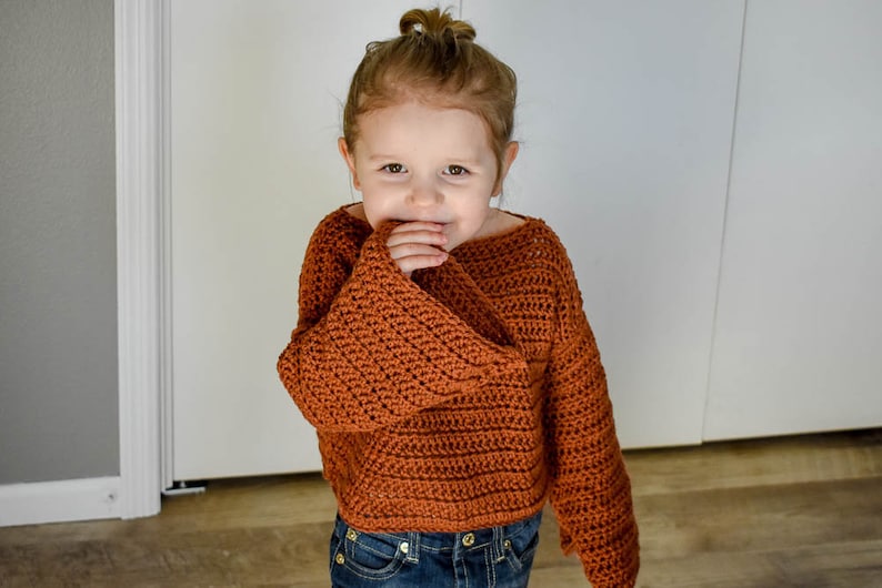 May include: A young child wearing a brown crocheted sweater with bell sleeves. The sweater has a loose fit and a slightly cropped length.