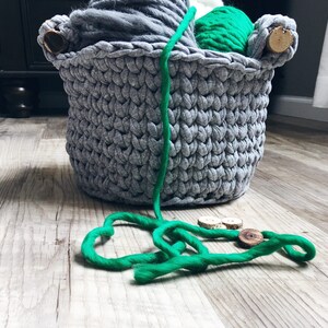May include: A gray crocheted basket filled with yarn in various shades of gray and green. The basket has wooden handles and is sitting on a wooden floor. A length of green yarn is lying on the floor in front of the basket.