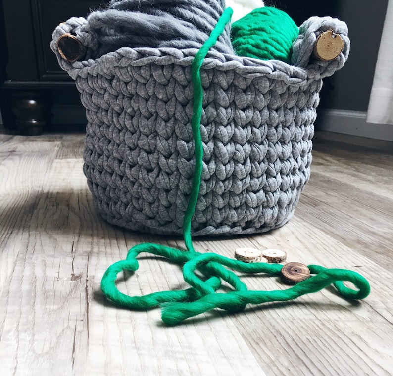 May include: A gray crocheted basket filled with yarn in various shades of gray and green. The basket has wooden handles and is sitting on a wooden floor. A length of green yarn is lying on the floor in front of the basket.