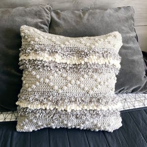 May include: A gray and white crocheted throw pillow with a textured surface and fringe trim. The pillow is on a gray and black bed.