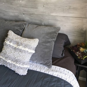 May include: A gray and white crocheted throw pillow with a textured surface sits on a gray bed with a white and gray geometric patterned bedspread. The bed is against a gray wood headboard.