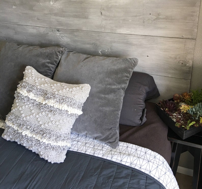 May include: A gray and white crocheted throw pillow with a textured surface sits on a gray bed with a white and gray geometric patterned bedspread. The bed is against a gray wood headboard.