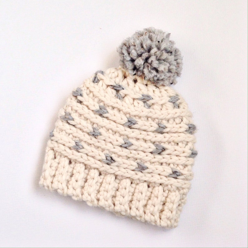 Puede incluir: Un gorro de punto blanco con un pomp&oacute;n gris y detalles grises. El gorro tiene un patr&oacute;n de punto texturizado.