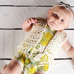 May include: A baby wearing a white crocheted vest with fringe and a lemon print onesie. The baby is lying on a white wooden surface.