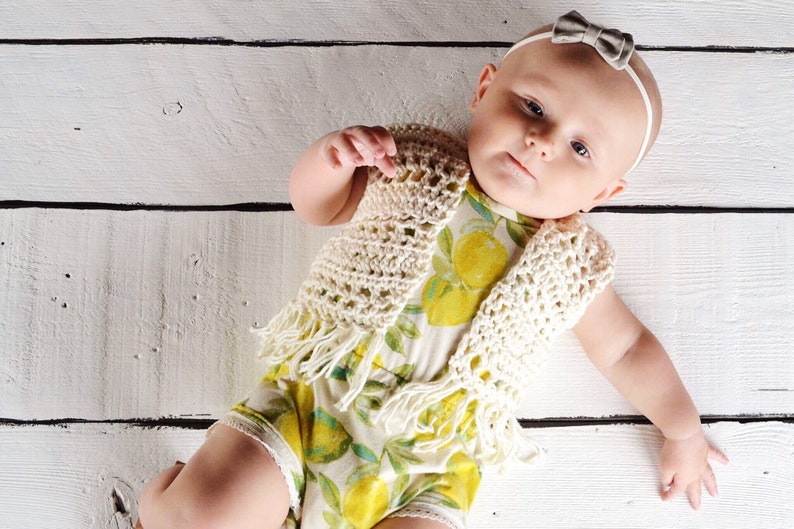 May include: A baby wearing a white crocheted vest with fringe and a lemon print onesie. The baby is lying on a white wooden surface.