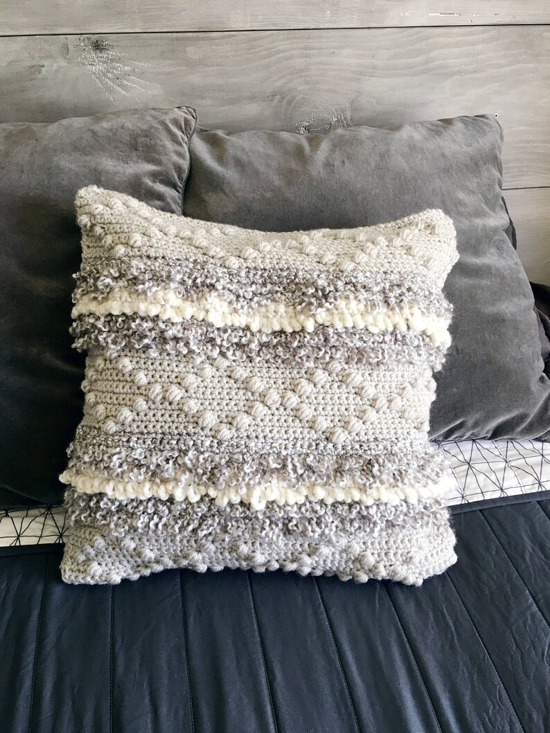 May include: A gray and white crocheted throw pillow with a textured surface and fringe trim. The pillow is on a gray and black bed.
