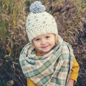 Puede incluir: Un ni&ntilde;o peque&ntilde;o con un gorro blanco de ganchillo con un pomp&oacute;n gris y una bufanda a cuadros. El ni&ntilde;o sonr&iacute;e y mira a la c&aacute;mara. El fondo es una imagen borrosa de hierba alta.