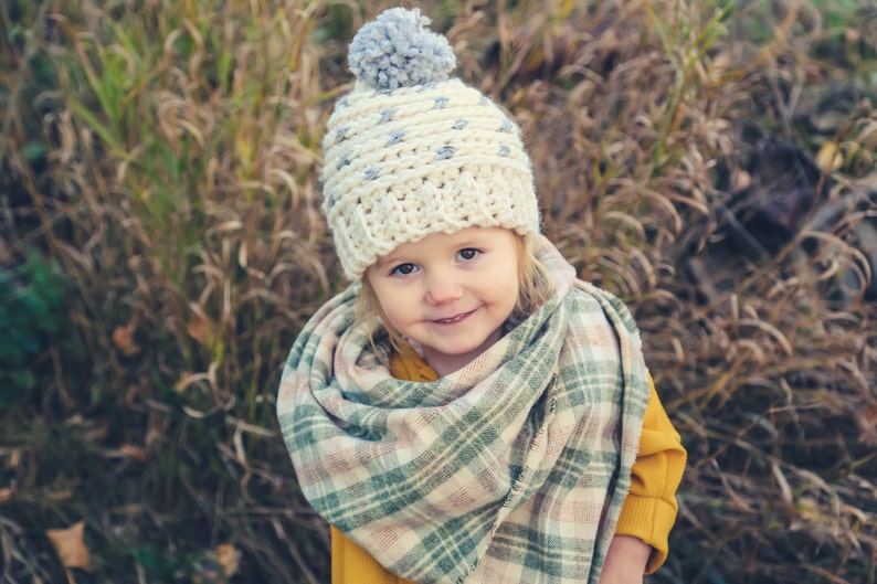 Puede incluir: Un ni&ntilde;o peque&ntilde;o con un gorro blanco de ganchillo con un pomp&oacute;n gris y una bufanda a cuadros. El ni&ntilde;o sonr&iacute;e y mira a la c&aacute;mara. El fondo es una imagen borrosa de hierba alta.