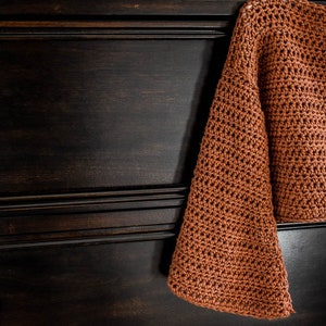 May include: A brown crocheted sweater with long sleeves and a cropped length. The sweater is hanging on a dark wooden dresser with two drawers.