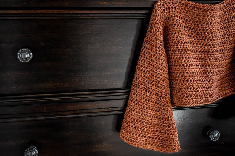 May include: A brown crocheted sweater with long sleeves and a cropped length. The sweater is hanging on a dark wooden dresser with two drawers.