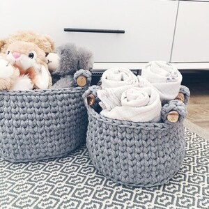 May include: Two gray crocheted baskets with wooden handles. One basket is filled with stuffed animals, including a bunny, a bear, and a gray cat. The other basket is filled with rolled up white towels.