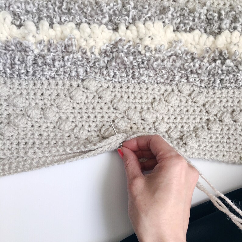 May include: A close-up of a hand using a needle to sew the edge of a crocheted blanket. The blanket is made with a textured yarn in shades of gray and white.