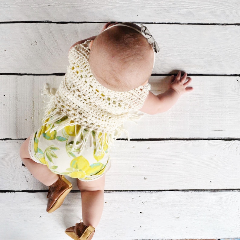 May include: A baby wearing a white crocheted top with fringe and a lemon print romper. The baby is crawling on a white wooden floor.