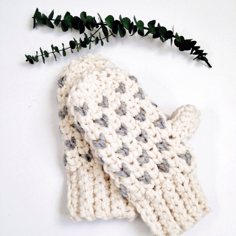 May include: A pair of white crocheted mittens with gray heart accents. The mittens are folded together and are lying on a white surface.