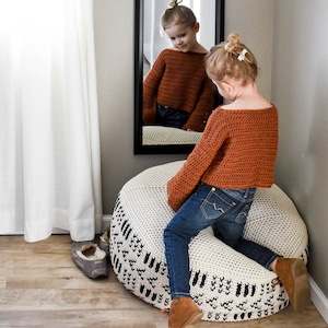 May include: A young girl wearing a brown crocheted sweater and blue jeans sits on a white and black crocheted pouf. The pouf has a geometric pattern.