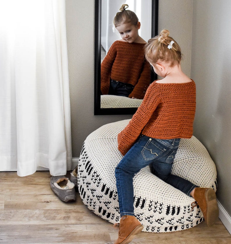 May include: A young girl wearing a brown crocheted sweater and blue jeans sits on a white and black crocheted pouf. The pouf has a geometric pattern.
