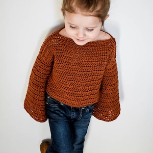 May include: A young child wearing a brown crocheted sweater with bell sleeves and blue jeans. The sweater has a loose fit and a slightly cropped length.