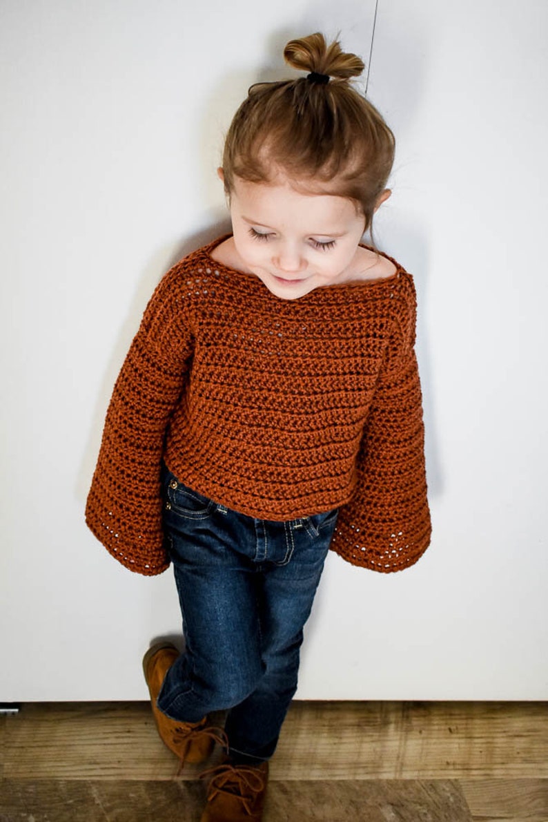 May include: A young child wearing a brown crocheted sweater with bell sleeves and blue jeans. The sweater has a loose fit and a slightly cropped length.