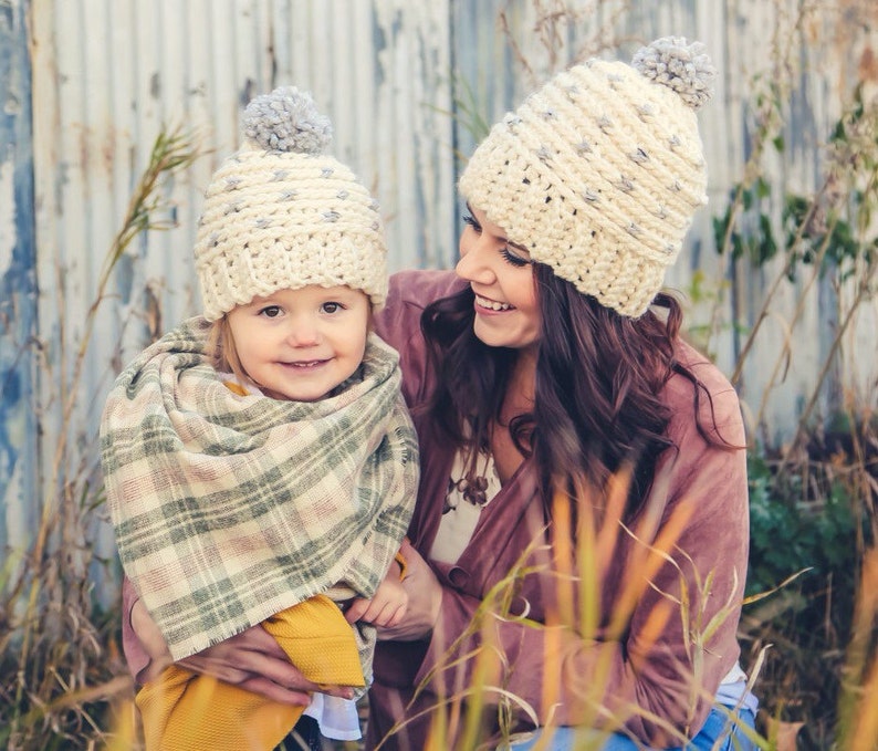 Puede incluir: Una mujer y una ni&ntilde;a con gorros de punto a juego con pompones grises. La mujer lleva un jersey granate y la ni&ntilde;a lleva una bufanda de cuadros y un jersey amarillo.