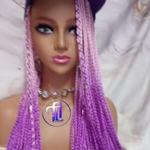 Pode incluir: Uma cabeça de manequim com cabelo comprido e trançado em tons de rosa e roxo, coberta por um boné de beisebol roxo. As tranças caem em cascata, mostrando um efeito gradiente. Um colar de prata com pedras transparentes está em volta do pescoço.