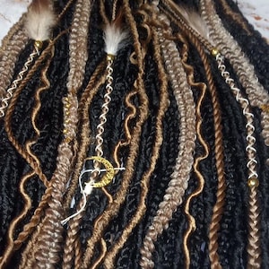 May include: A close-up of a bundle of synthetic dreadlocks in black, brown, and blonde colors. The dreadlocks have gold and white beads and charms.