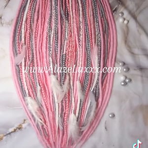 May include: Pink, white and grey synthetic dreadlocks with white feathers. The dreadlocks are tied together at the top.