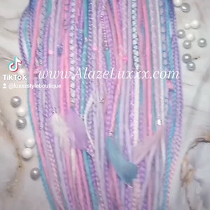 May include: A collection of colorful synthetic hair extensions in shades of pink, purple, blue, and white. The extensions are braided and have a variety of textures. The text "www.AlazeLuxxx.com" is visible in the image.