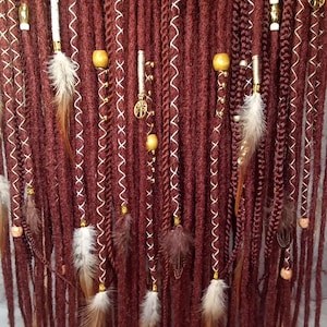 Peut inclure: Un ensemble de longues dreadlocks marrons avec des perles dorées et blanches, des plumes et des breloques. Les dreadlocks sont disposées en rangée et pendent.