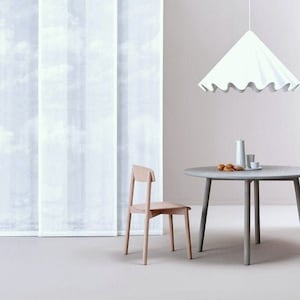 Könnte beinhalten: Minimalistisches Interieur mit einem runden grauen Tisch, einem hellen Holzstuhl und einer weißen Pendelleuchte mit gewelltem Rand. Im Hintergrund ein weißer Vorhang mit Wolkenmuster. Auf dem Tisch befinden sich kleine dekorative Gegenstände.