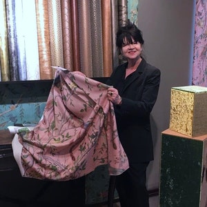 May include: A woman in a black blazer holds up a pink fabric with a floral pattern. The fabric has a light pink background with a delicate floral design in shades of green, brown, and white. The fabric is displayed in front of a wall with a variety of wallpaper samples.
