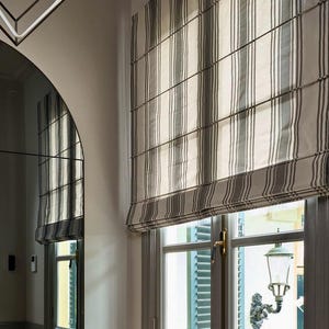 Puede incluir: Vista interior de una habitación con una ventana con una cortina romana a rayas. Una silla vintage con detalles ornamentados se encuentra frente a un gran espejo. La habitación tiene pisos de madera y un radiador. Botas negras cerca de la ventana.