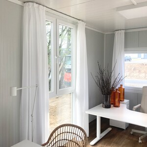 Peut inclure: Un bureau blanc avec une grande fen&ecirc;tre, des rideaux blancs, un bureau blanc, une chaise en osier et un vase avec des branches s&eacute;ch&eacute;es.