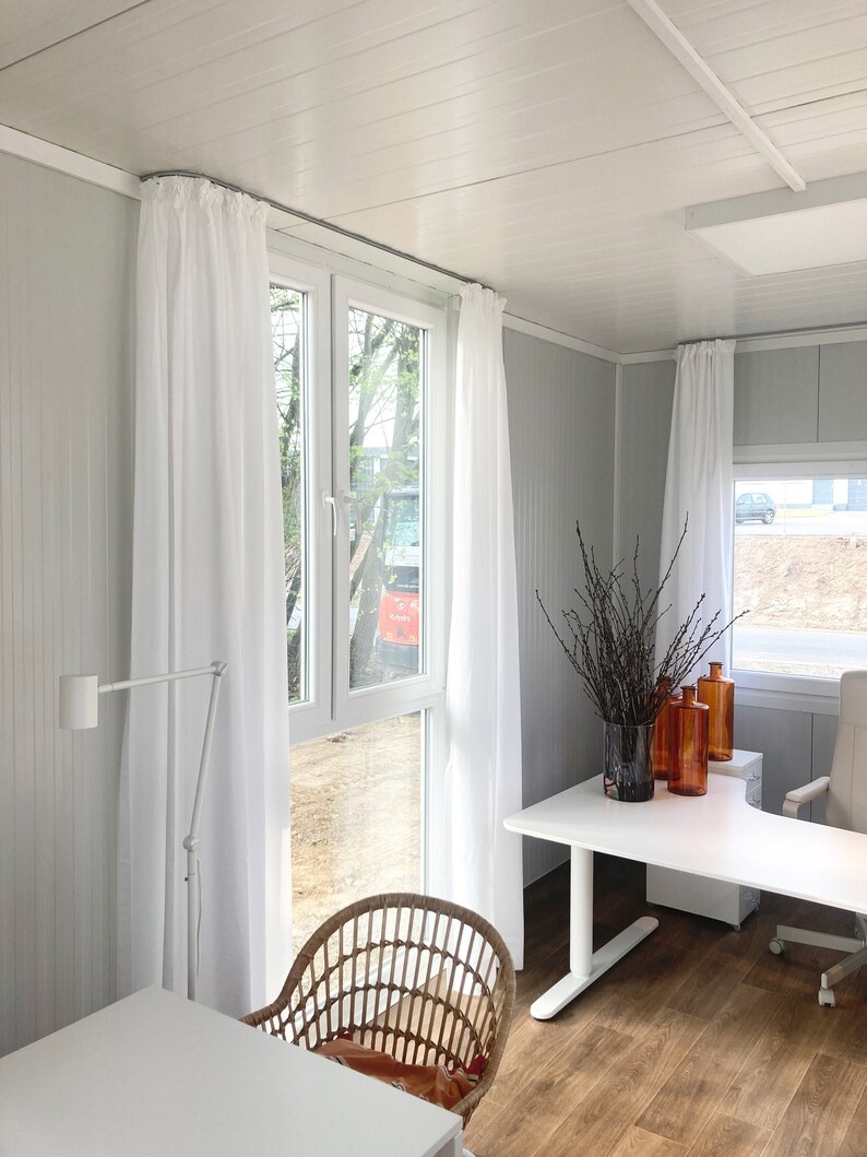 Peut inclure: Un bureau blanc avec un bureau blanc et une chaise en osier. Le bureau a une lampe blanche, un vase avec des branches et trois bouteilles en verre brun. La pi&egrave;ce a des rideaux blancs et une grande fen&ecirc;tre avec vue sur les arbres.